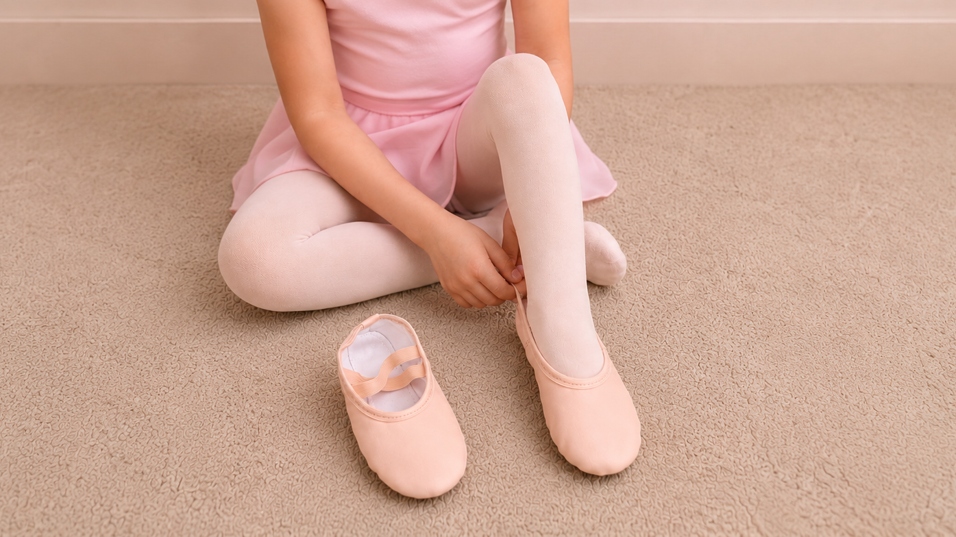 Child in pink ballet attire adjusting a ballet slipper on a neutral beige carpet before dance class.