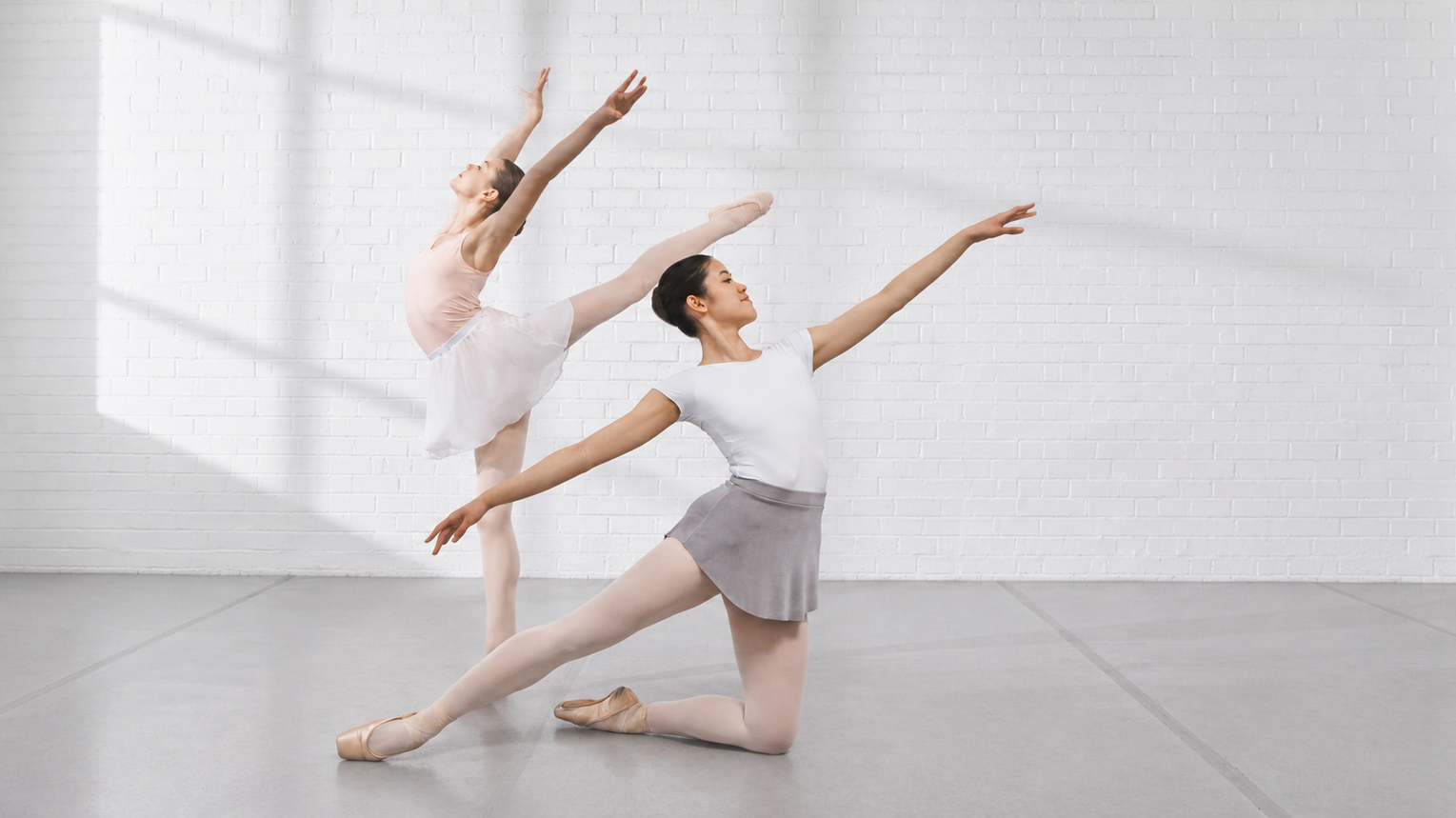 Two ballet dancers practicing in a bright studio, one kneeling in a gray skirt and white leotard while another extends into an arabesque against a white brick wall.