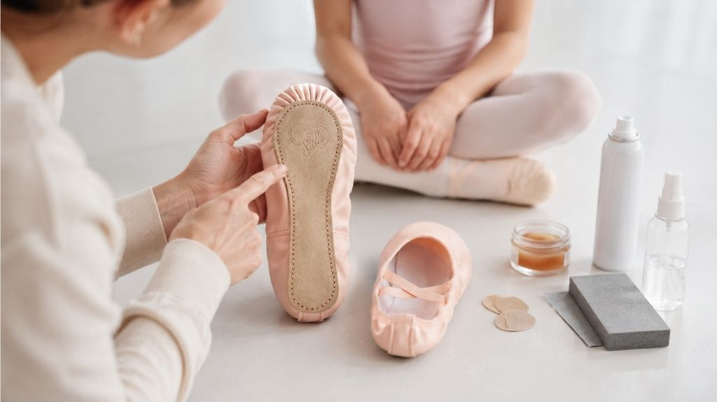 Adult hands showing the full suede sole of a pale pink ballet shoe beside rosin, grip pads, sandpaper, and spray bottles, with a young dancer seated in the background.