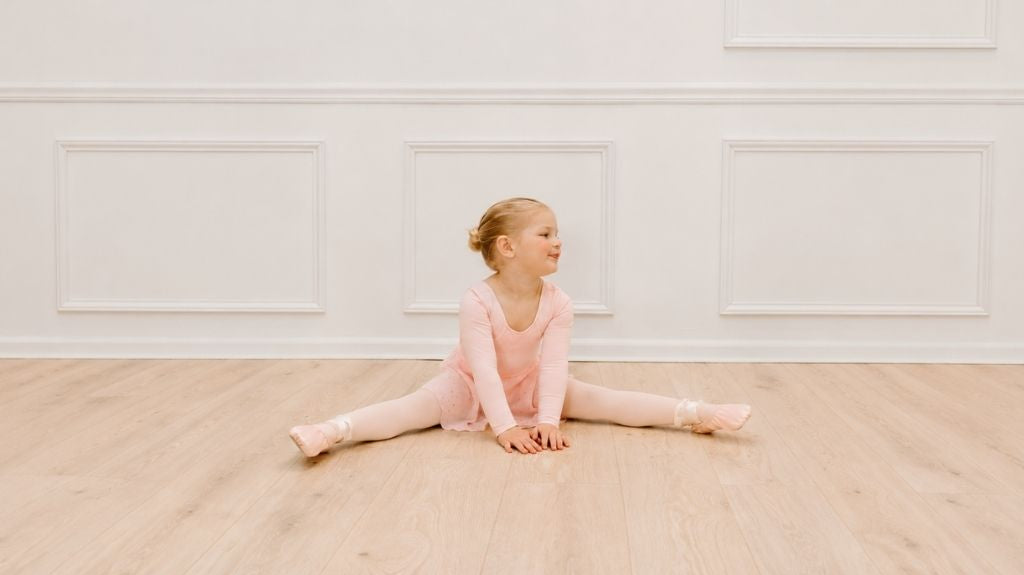 Toddler ballerina in a pink long-sleeve leotard, tights, and ballet shoes sitting in a split stretch on a light wood floor in a bright studio.