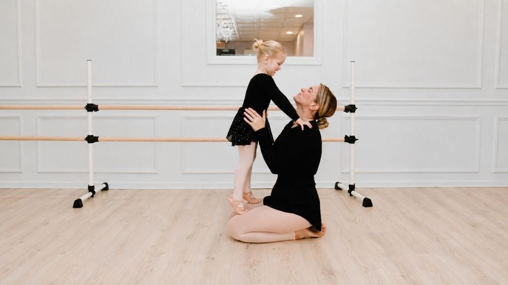 Young ballet student in a black leotard smiling with her teacher during a gentle class moment in a bright dance studio.