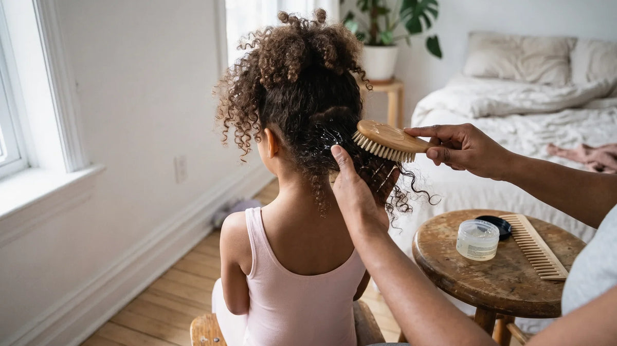 Parent smoothing gel into a girl's natural curls with a bristle brush to start a ballet bun on curly hair