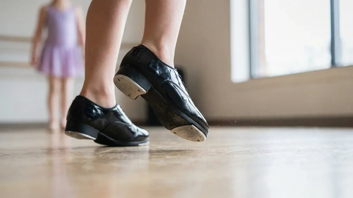 What is tap dance: a young dancer's black tap shoes tapping out a rhythm on a light wood studio floor