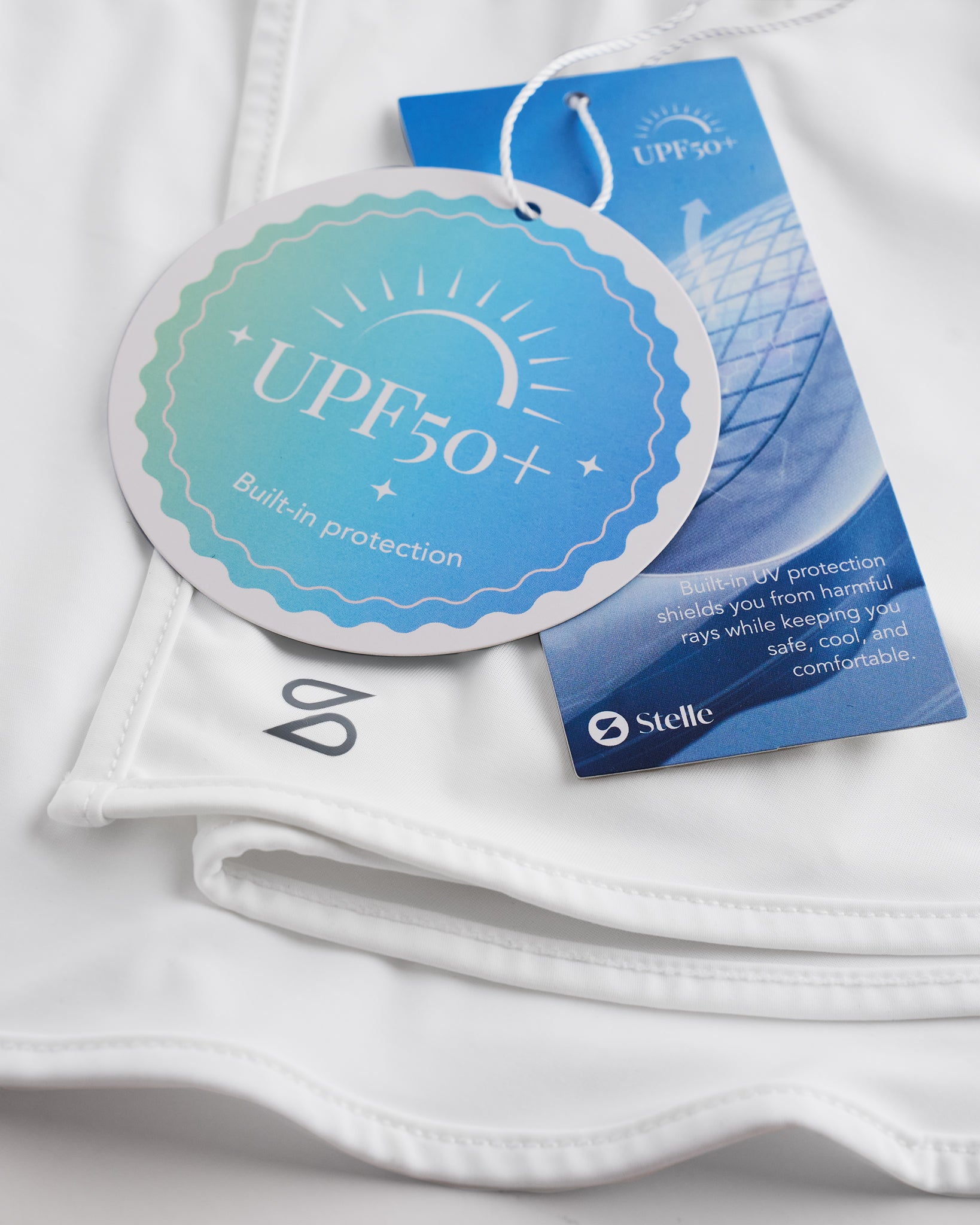 Close-up of white sunscreen face mask showing UPF 50+ built-in protection tag, smooth fabric, and Stelle logo.