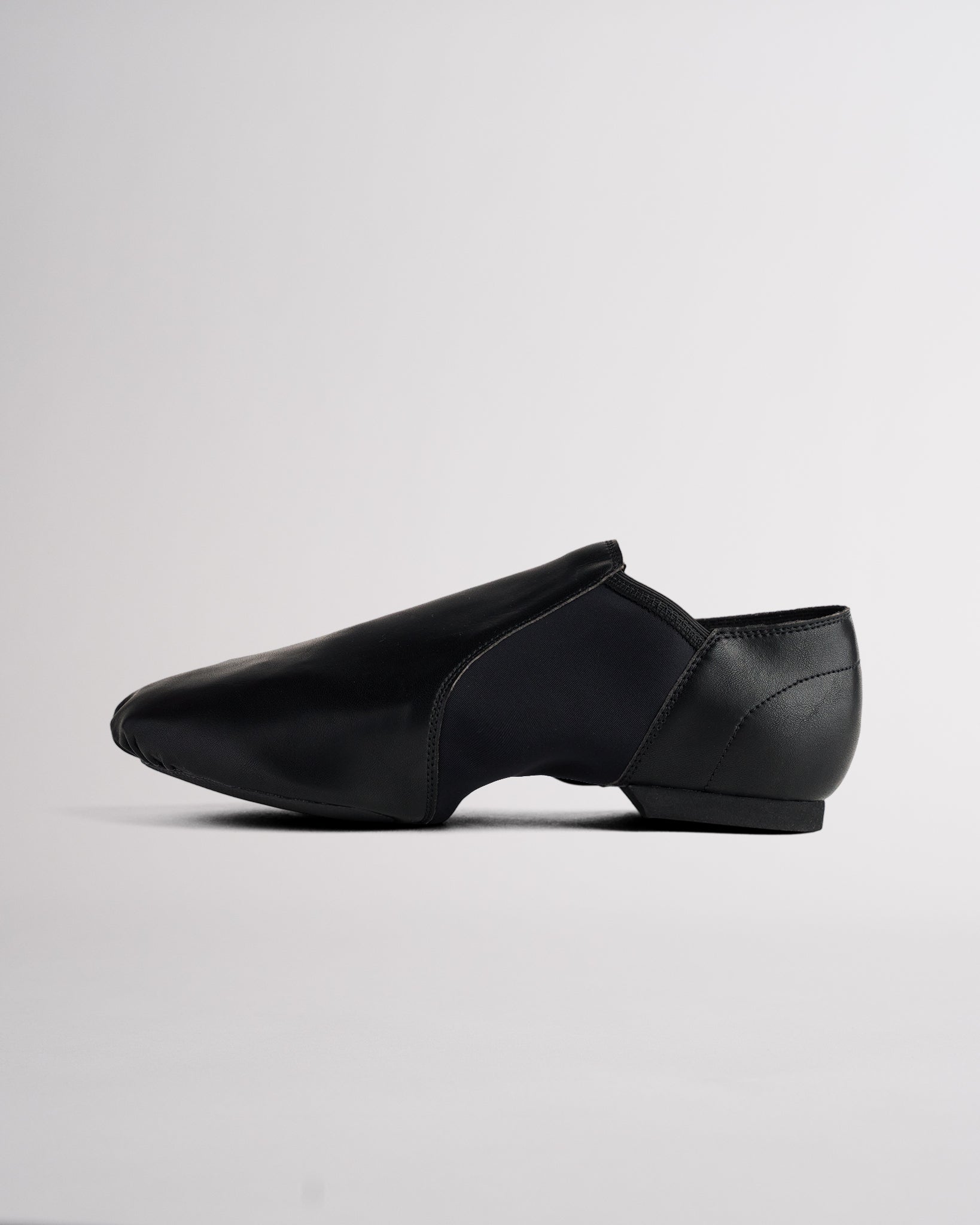 Side view of black vegan leather jazz shoe showing slip-on design, stitched details, and flexible dance shoe shape.