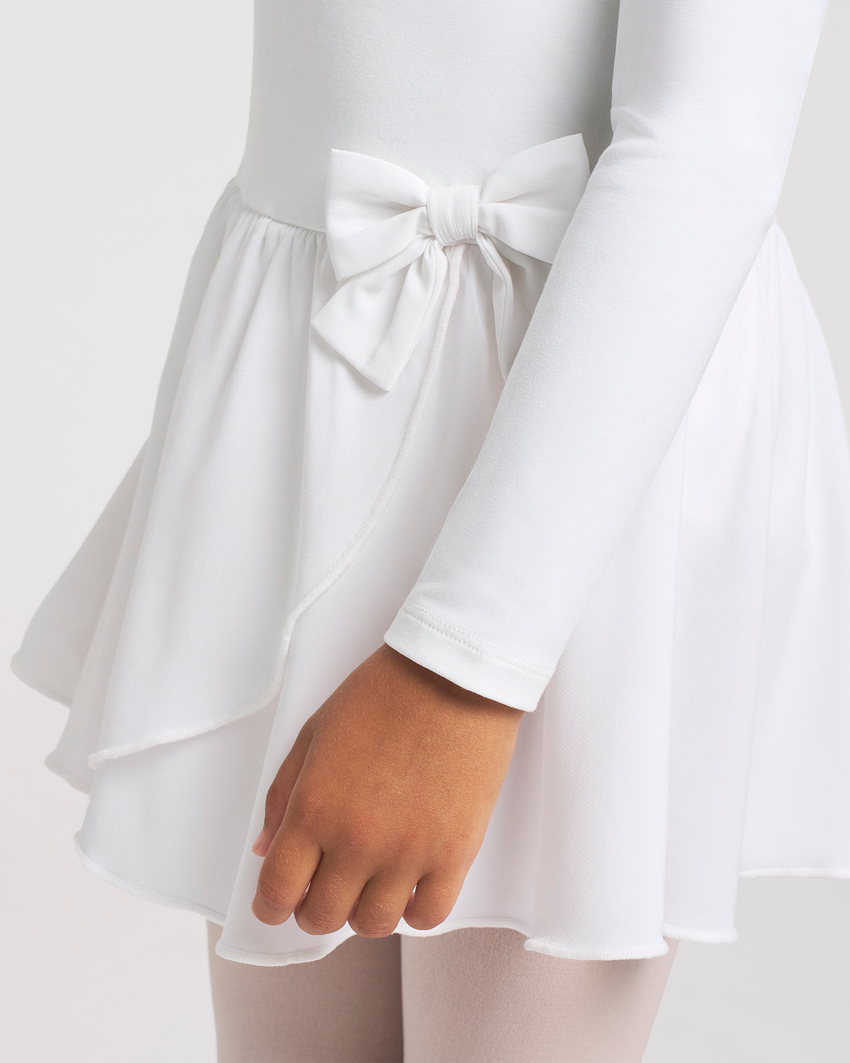 Close-up of white girls dance dress showing the bow accent, layered chiffon wrap skirt, and smooth long sleeve bodice.
