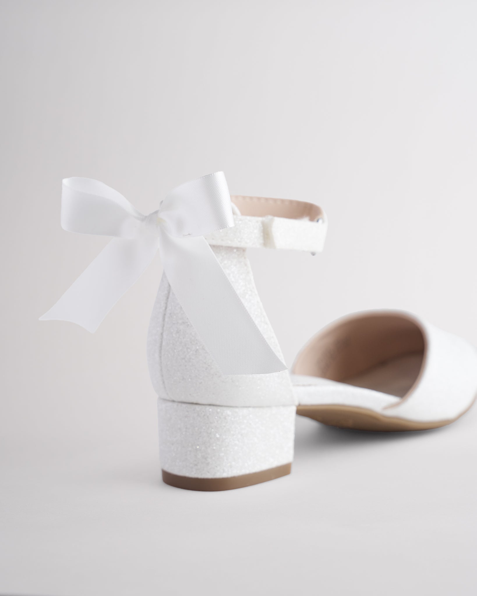 Girls’ white glitter low block heel dress shoes with ankle straps and removable satin bow accessories.