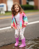 Child wearing a colorful raincoat and pink boots standing in a puddle on a street.