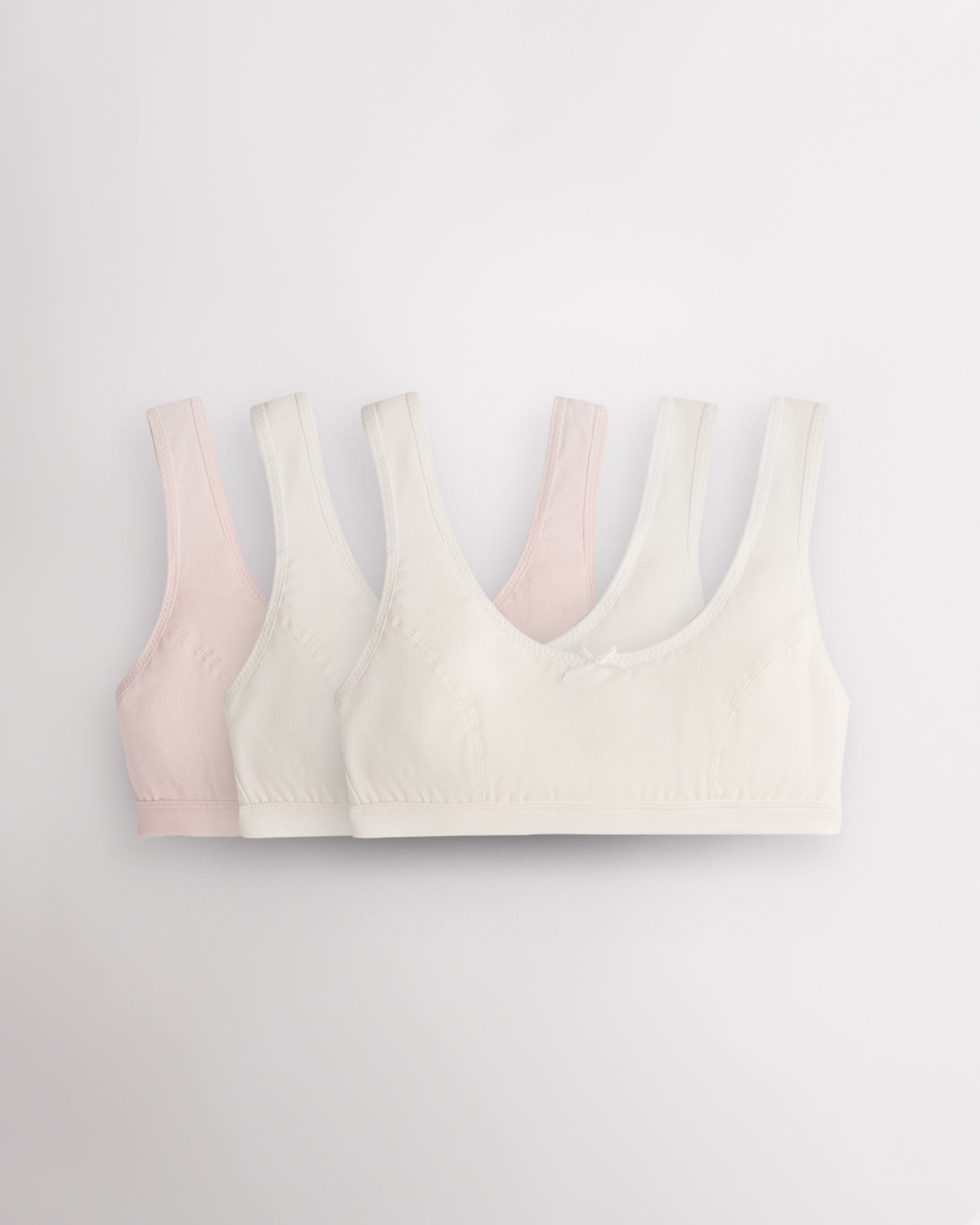 Three Stelle girls cotton bralettes in white and whisper pink with wide straps, soft lining, and small bow detail.