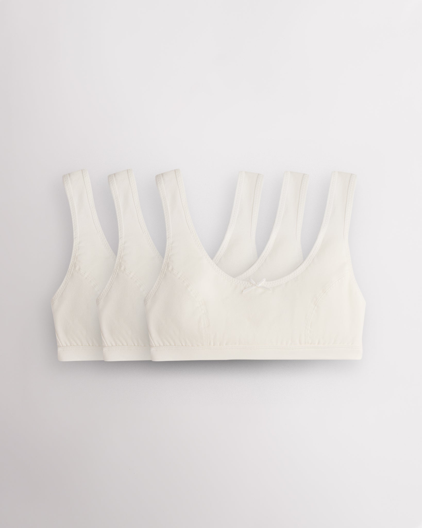 Three Stelle girls cotton bralettes in white with wide straps, soft lining, and small bow detail.