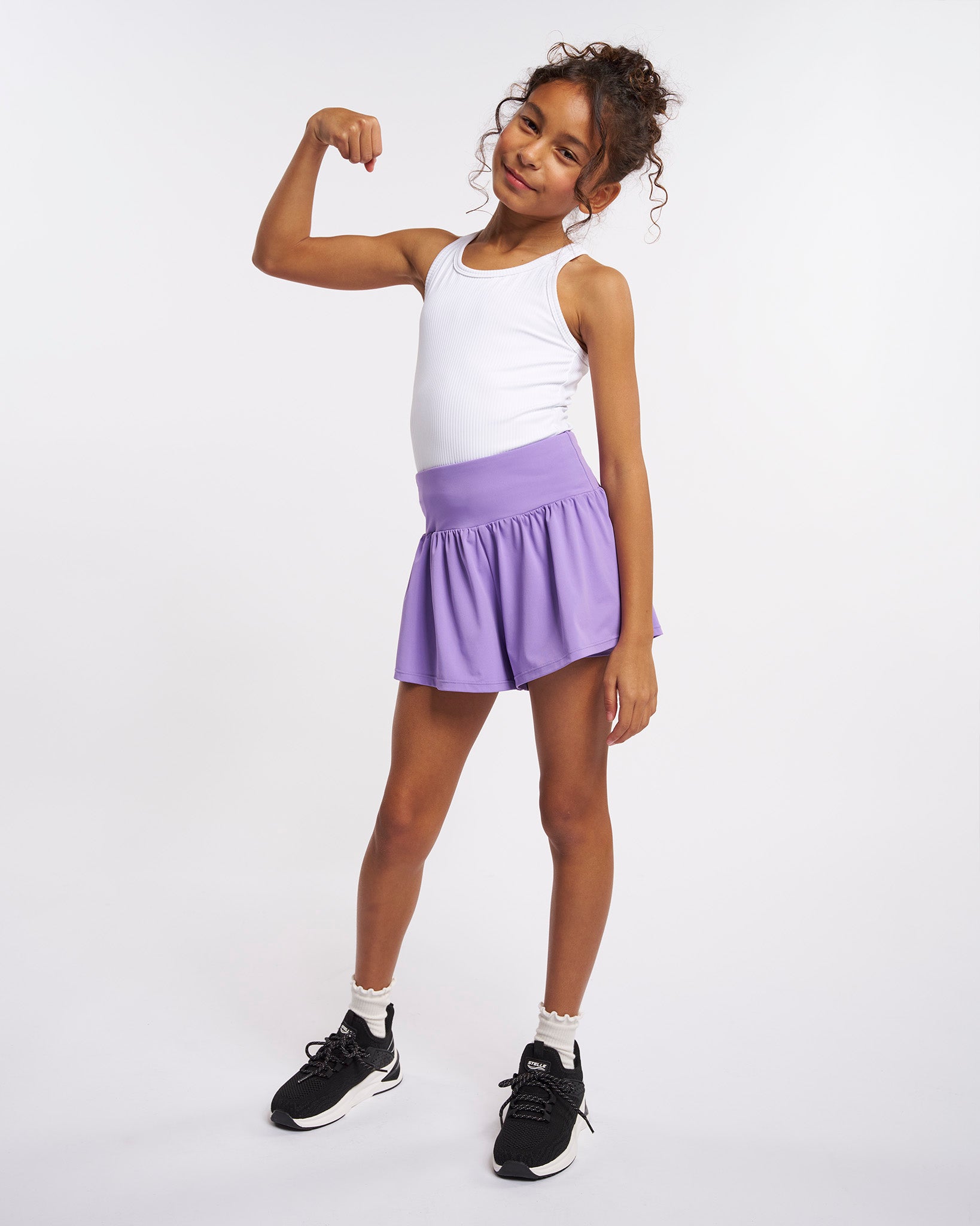 Girl wearing lavender high waisted 2-in-1 flutter shorts with built-in liner and white tank top, posing and flexing arm