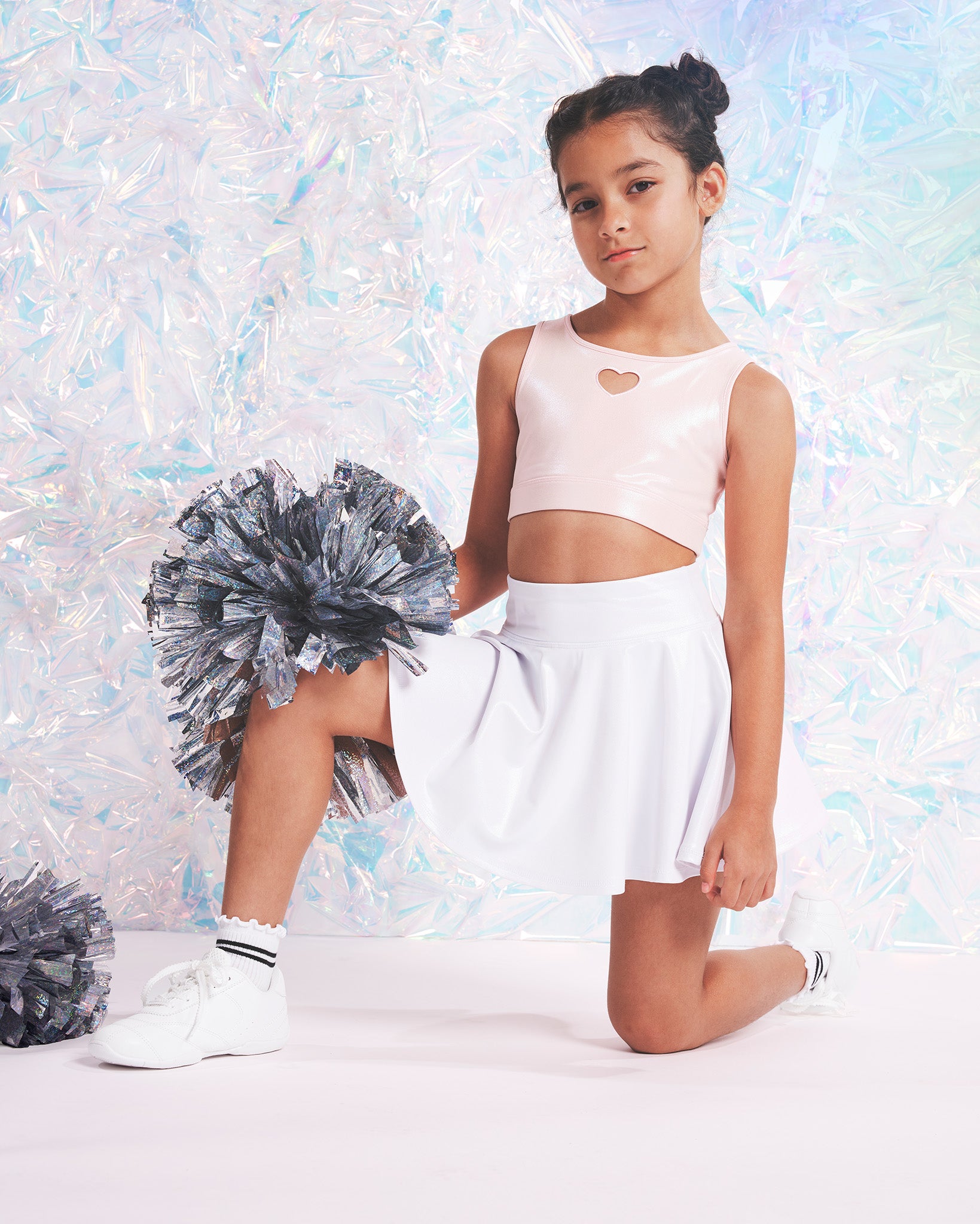 Girl wearing a shimmer heart crop top, white A-line skirt, kids socks, and white dance shoes while holding silver pom-poms.