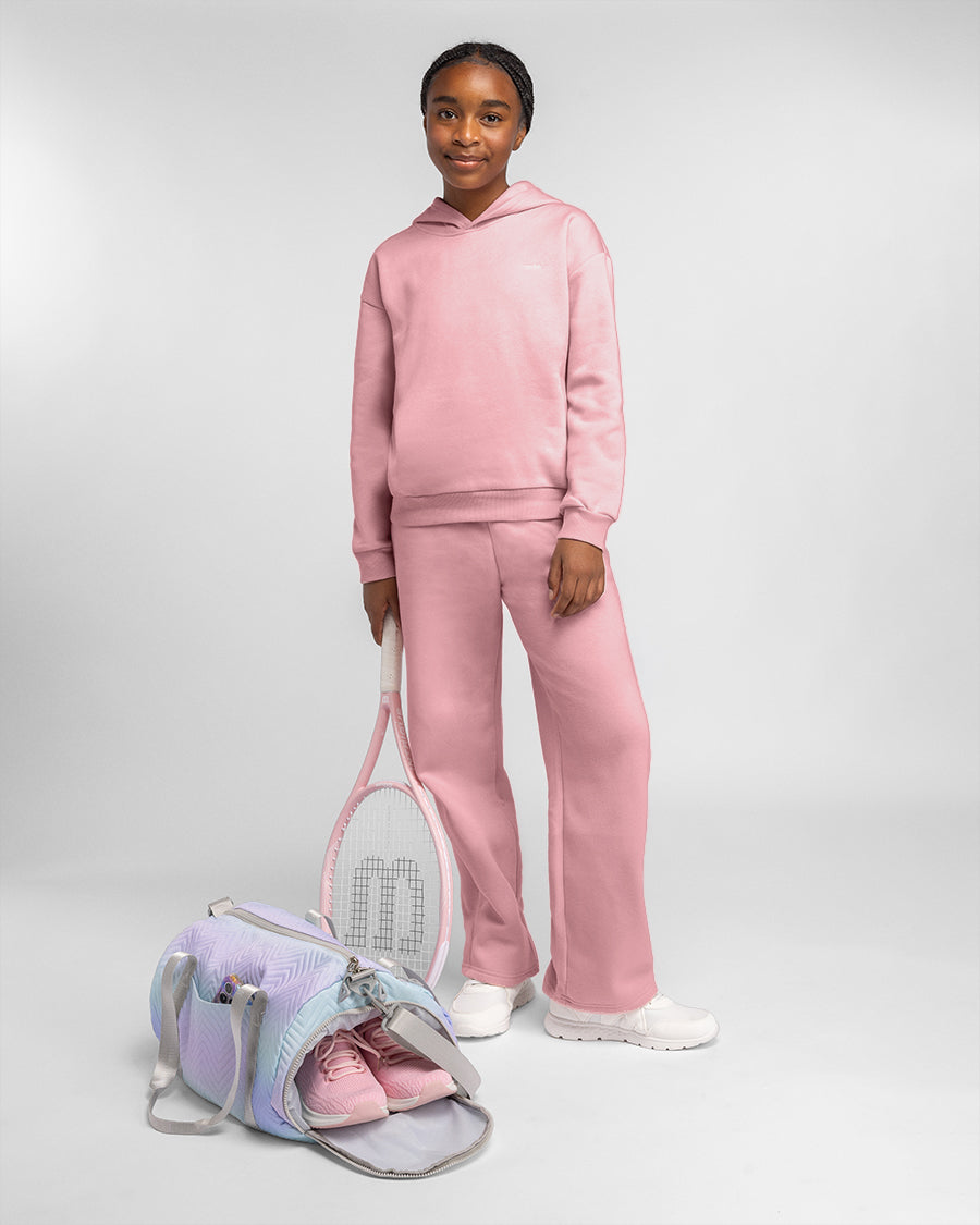 Girl wearing a matching pink sweatshirt and fleece sweatpants set with white sneakers beside a pastel duffel bag.