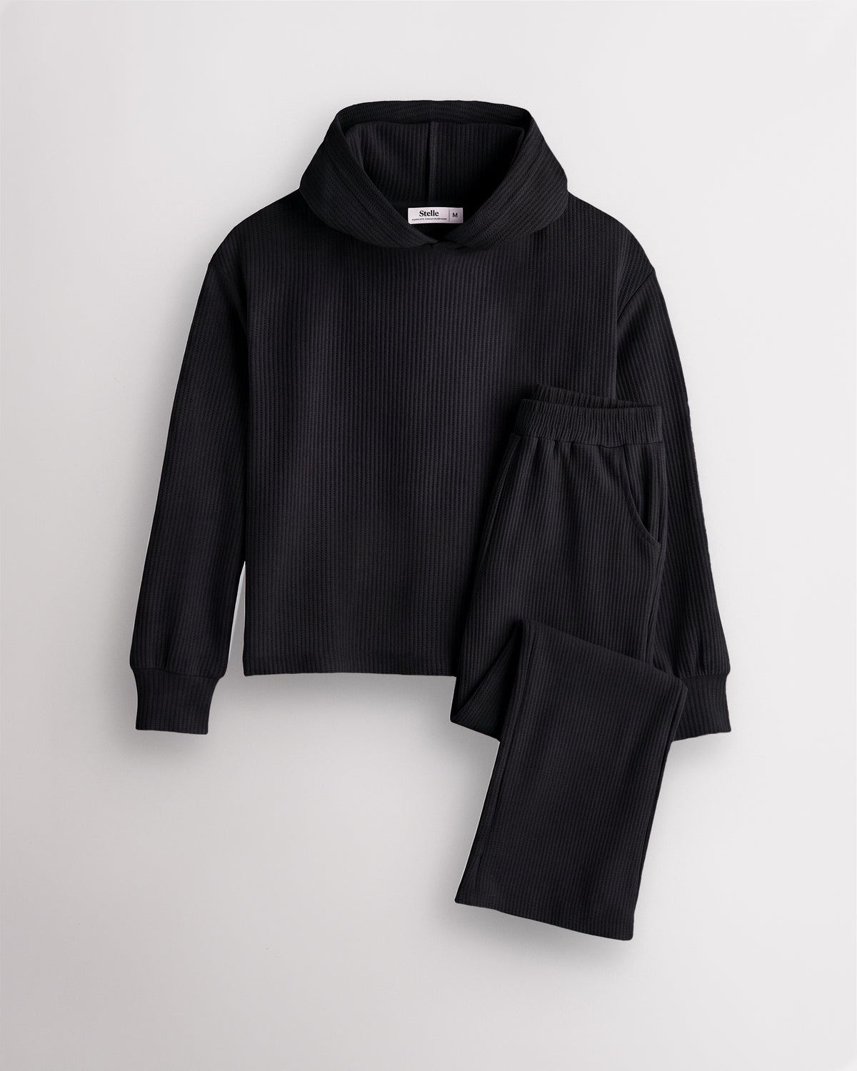 Black Stelle kids waffle knit lounge set with a hooded top and matching pants, laid flat on a grey background.