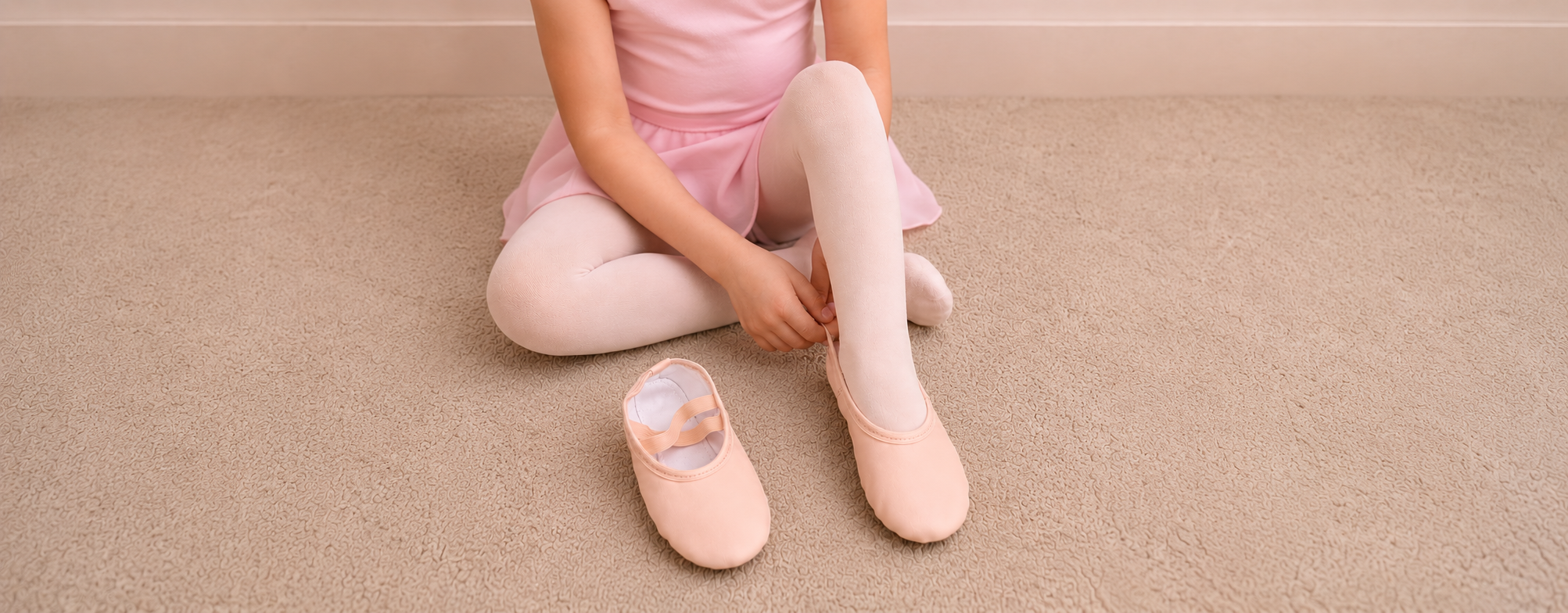 Child in pink ballet attire adjusting a ballet slipper on a neutral beige carpet before dance class.