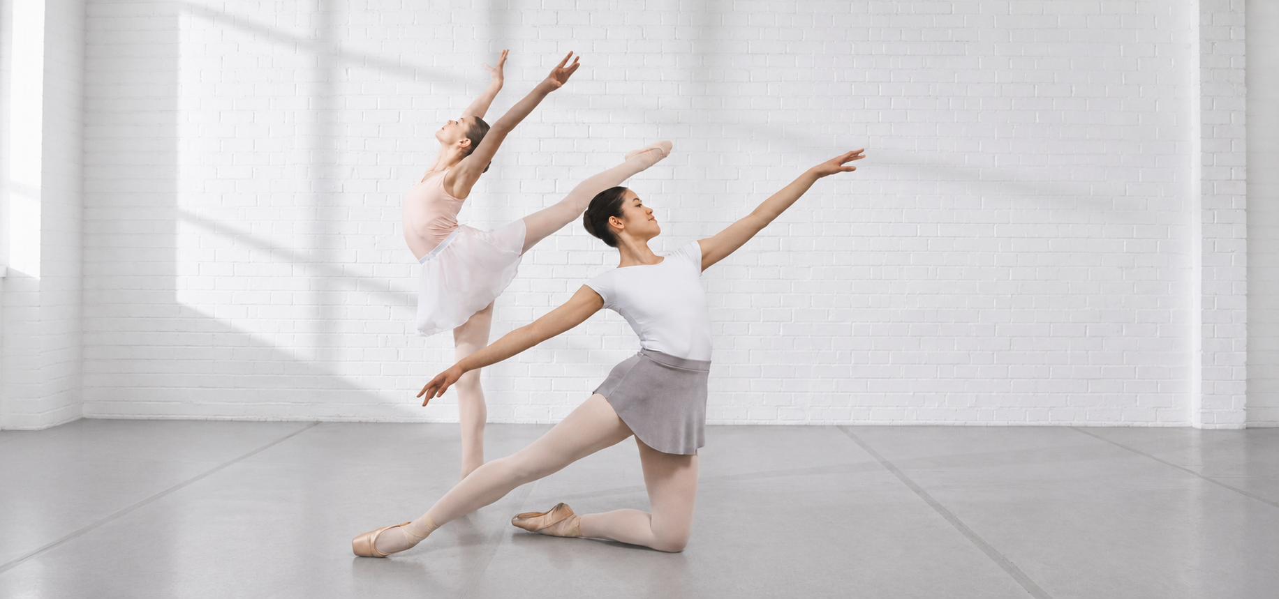 Two ballet dancers practicing in a bright studio, one kneeling in a gray skirt and white leotard while another extends into an arabesque against a white brick wall.
