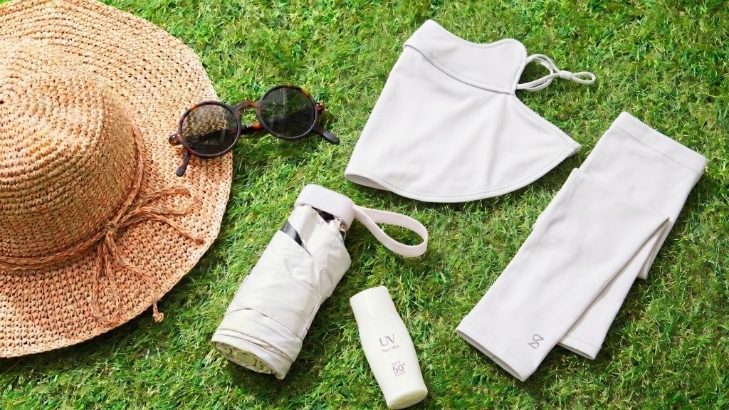 Beige UV protection accessories arranged on grass, including a wide-brim straw hat, sunglasses, face mask, arm sleeves, folded umbrella, and sunscreen bottle.