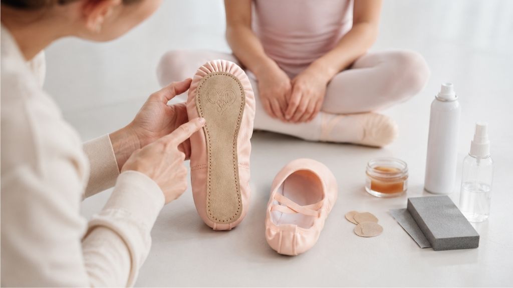 Adult hands showing the full suede sole of a pale pink ballet shoe beside rosin, grip pads, sandpaper, and spray bottles, with a young dancer seated in the background.