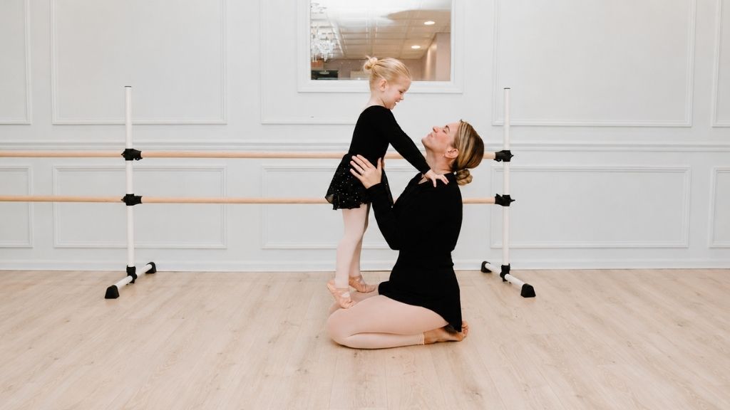 Young ballet student in a black leotard smiling with her teacher during a gentle class moment in a bright dance studio.