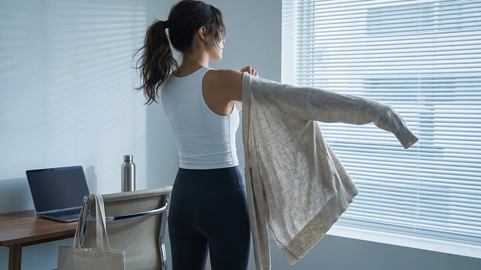 Woman pulling a lightweight cardigan over a summer athleisure outfit at her office desk, the layering fix for indoor AC