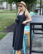 Woman in a long black dress and hat holding a cup, standing outdoors near tables and chairs.