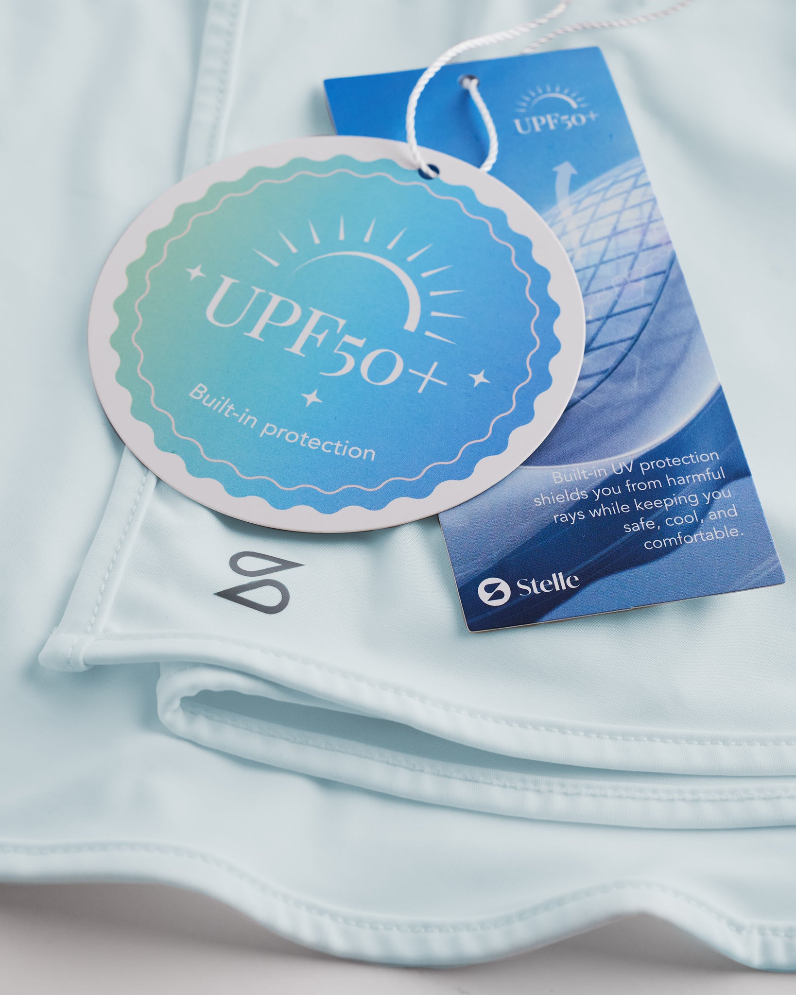 Close-up of Light Blue sunscreen face mask showing UPF 50+ built-in protection tag, smooth fabric, and Stelle logo.