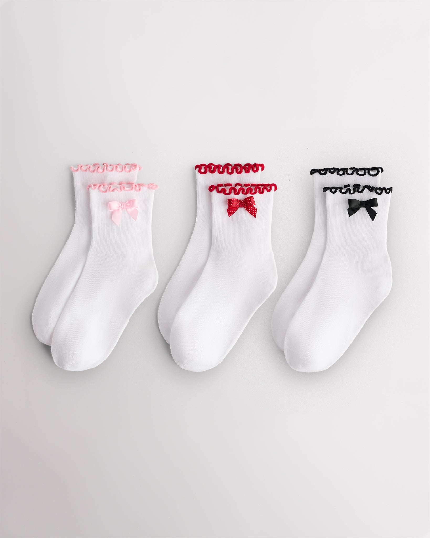 Three pairs of white kids socks with pink, red, and black ribbon bows and matching ruffle cuffs