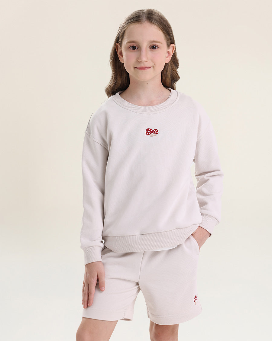 Young girl wearing a light gray sweatshirt and shorts set with a small red logo on a beige background