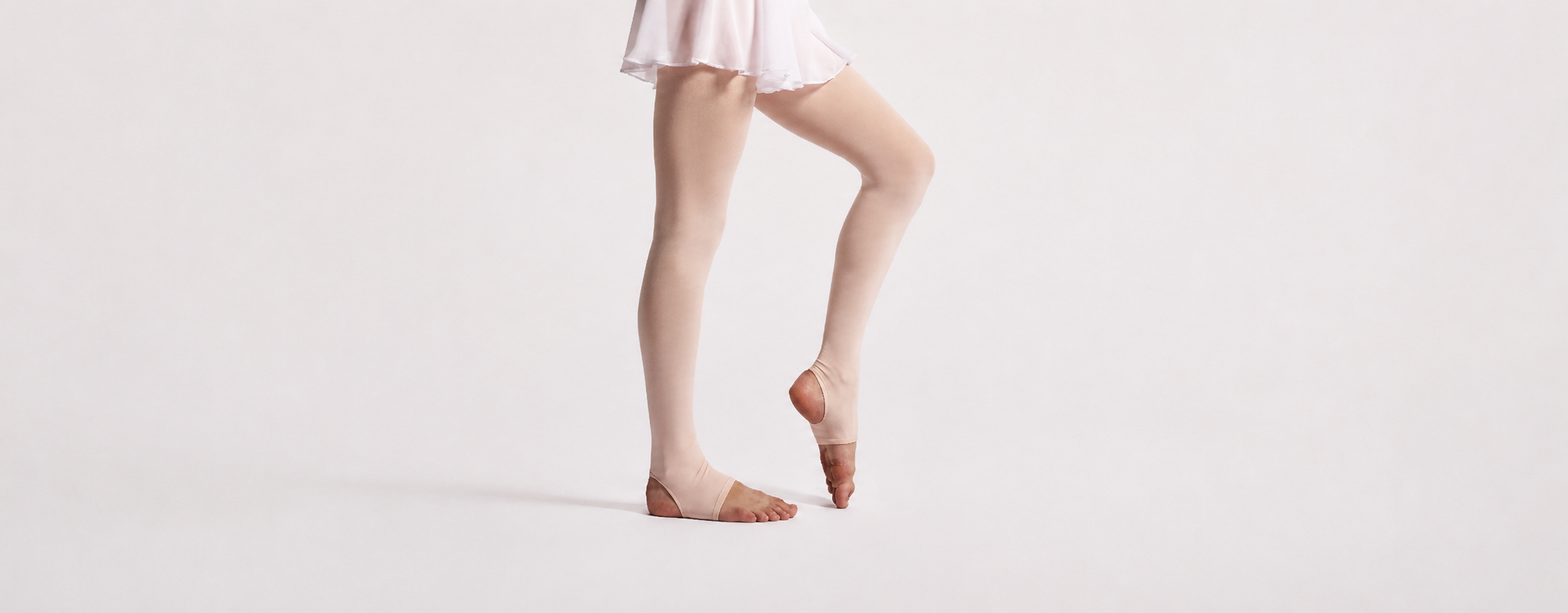 Dancer wearing light pink convertible ballet tights and a white ballet skirt, standing in a soft studio pose against a neutral background.