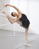 Young ballerina stretching at a barre in a black leotard and black mesh ballet skirt with pink tights and ballet slippers.