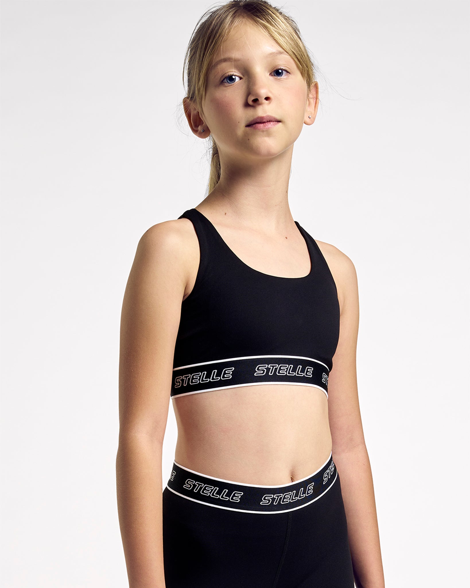 Girl wearing the black Stelle girls sports bra with a white logo underband, front view.