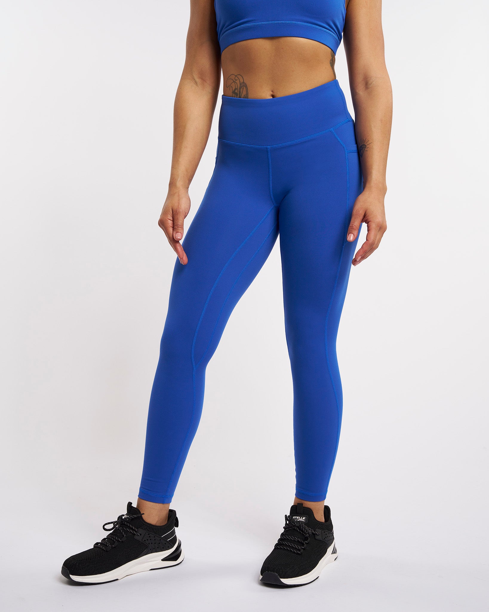 Blue high-waisted leggings with side pockets, contour seam details, and full-length fit styled with black sneakers.