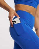 Close-up of blue leggings showing roomy side pocket holding a phone, high waistband, and curved seam detail.