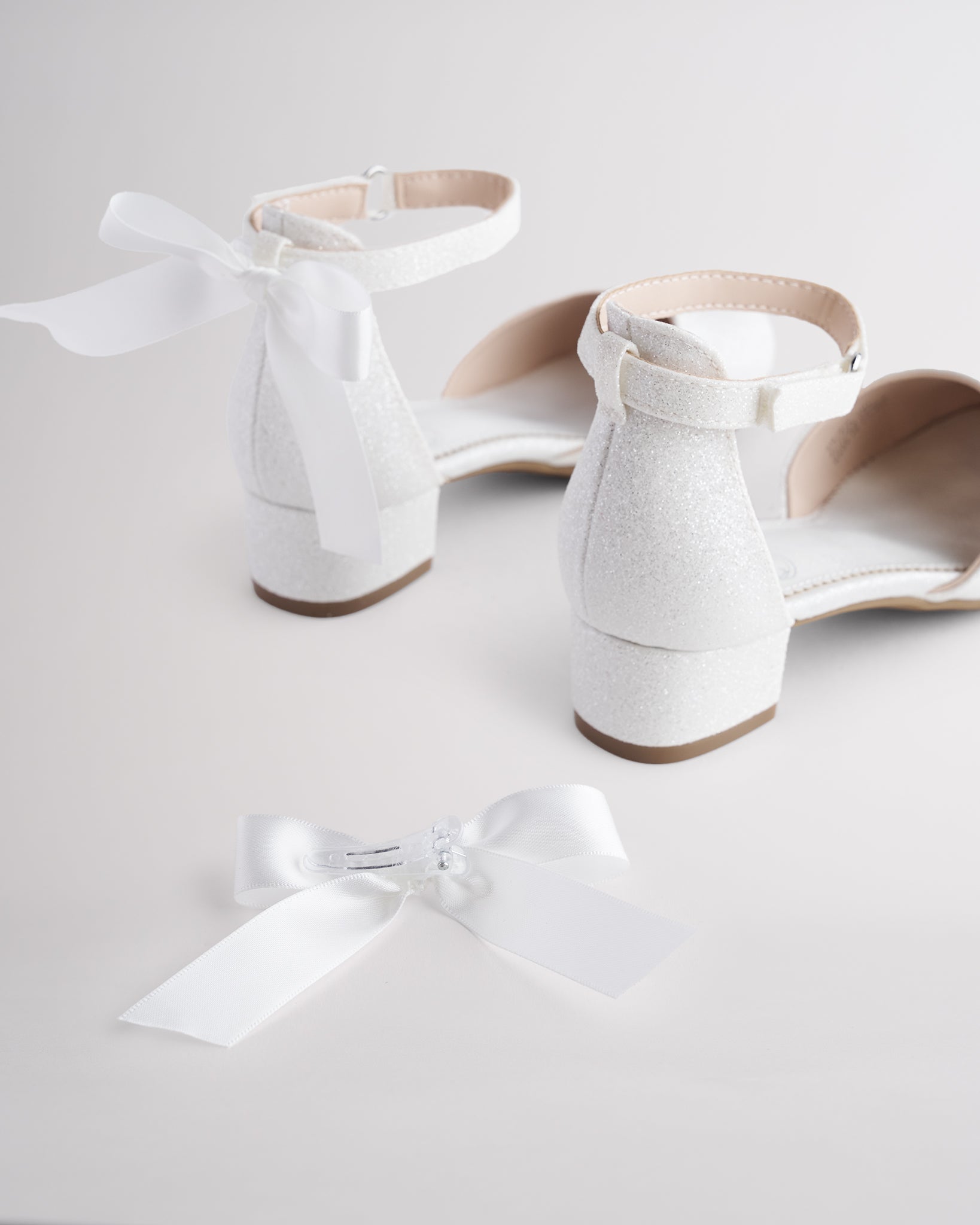 Girls’ white glitter low block heel dress shoes with ankle straps and removable satin bow accessories.