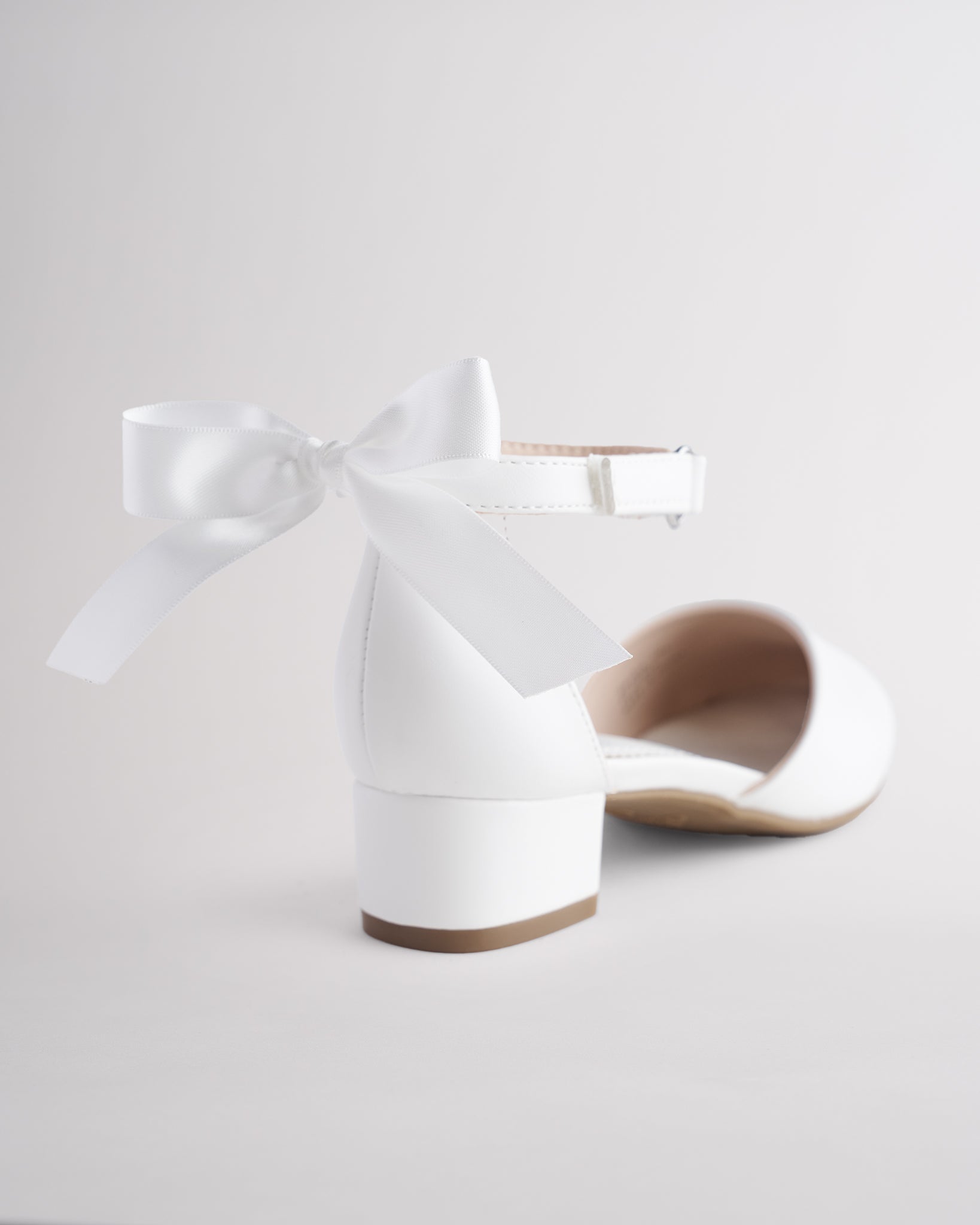 Girls’ white low block heel dress shoes with ankle straps and removable satin bow accessories.