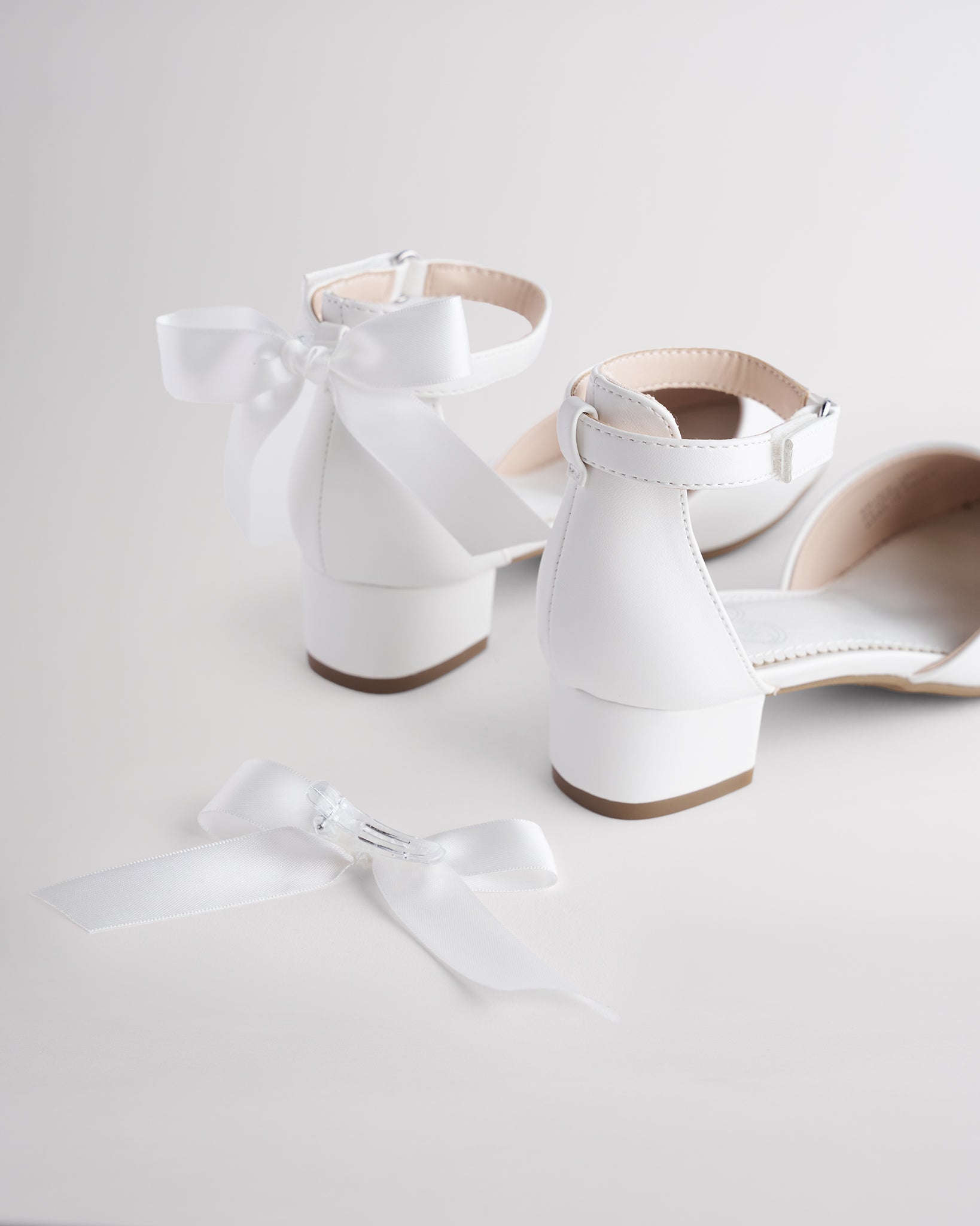 Girls’ white low block heel dress shoes with ankle straps and removable satin bow accessories.