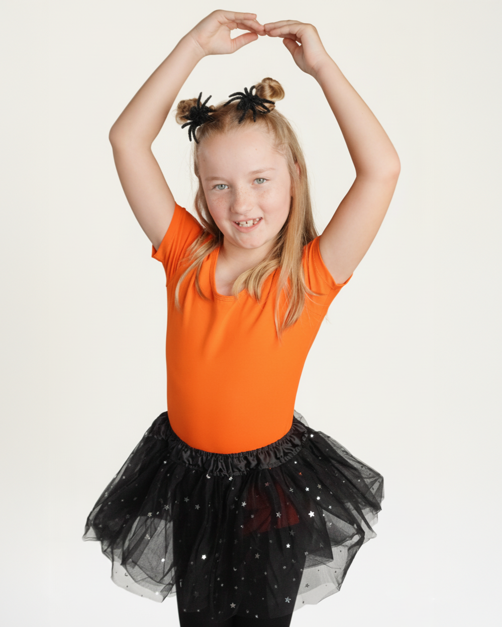 Young girl in an orange top and black tutu posing with arms raised against a white background