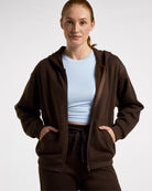 Woman wearing the brown Stelle fleece-lined zip-up hoodie in Coffee, front view with hands in the kangaroo pockets.