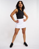 Full-body view of a woman wearing a black cropped polo tank top with a white athletic skort and black sneakers.