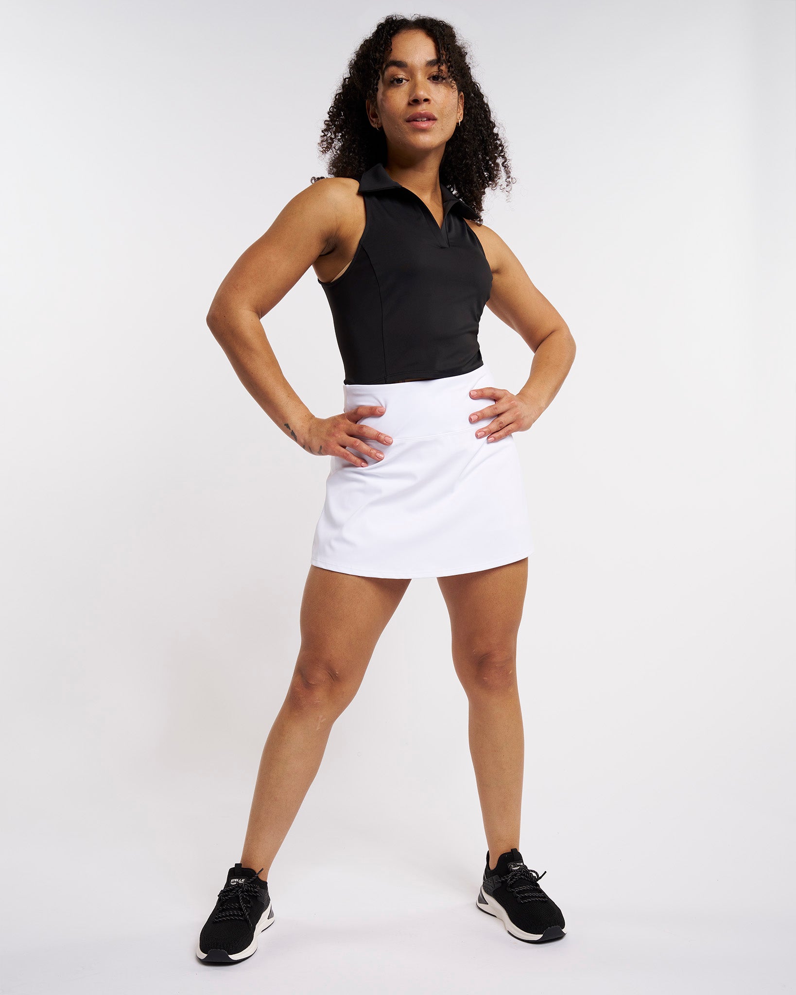 Full-body view of a woman wearing a black cropped polo tank top with a white athletic skort and black sneakers.