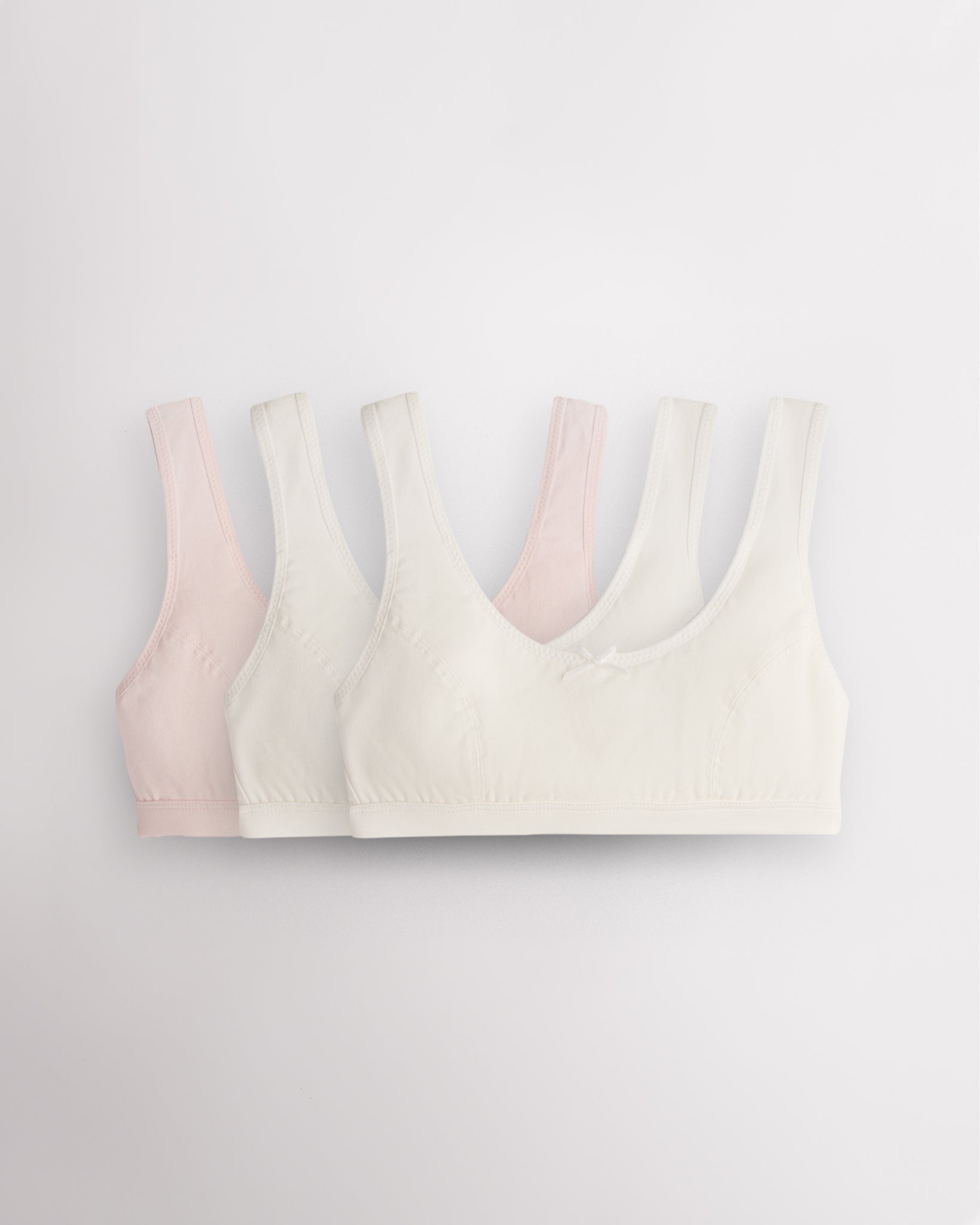 Three Stelle girls cotton bralettes in white and whisper pink with wide straps, soft lining, and small bow detail.
