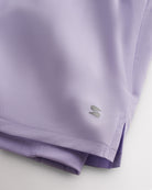 Close-up of lavender Stelle running shorts showing smooth fabric, subtle side slit, inner shorts liner, and logo detail.