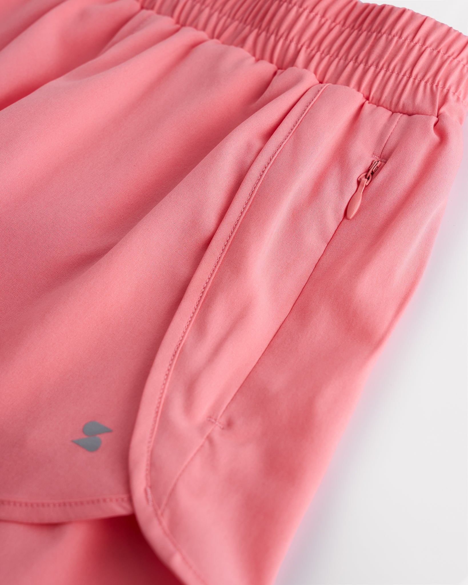 Close-up of girls coral running shorts showing elastic waistband, curved seam, and zipper pocket detail.