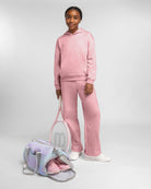 Girl wearing a matching pink sweatshirt and fleece sweatpants set with white sneakers beside a pastel duffel bag.