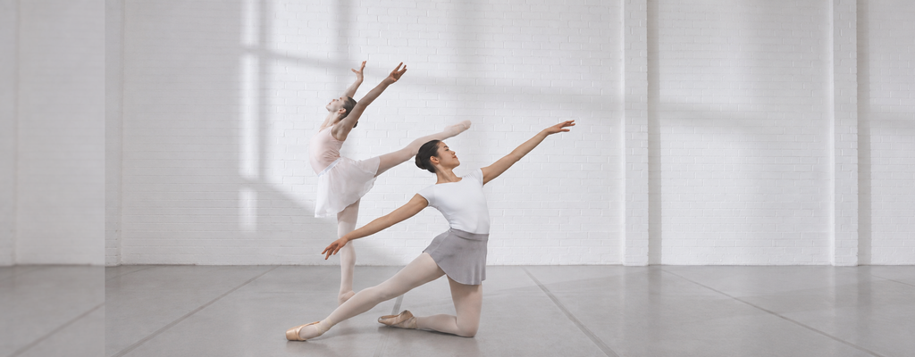 Two ballet dancers practicing in a bright studio, one kneeling in a gray skirt and white leotard while another extends into an arabesque against a white brick wall.