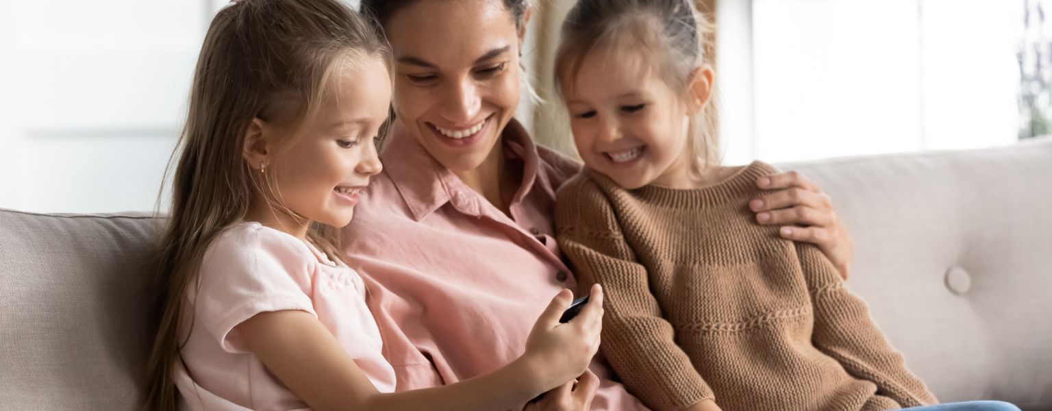 Mother sitting on a couch with two young daughters, smiling together while looking at a phone.