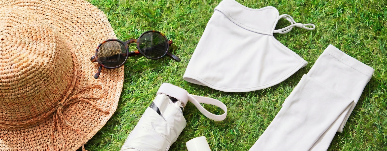 Beige UV protection accessories arranged on grass, including a wide-brim straw hat, sunglasses, face mask, arm sleeves, folded umbrella, and sunscreen bottle.