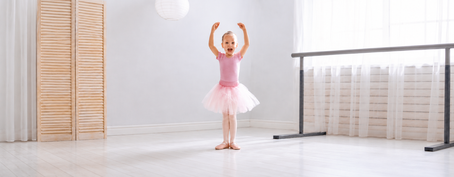Young girl in a pink ballet leotard, tutu, tights, and leather ballet shoes posing with arms raised in a bright dance studio.