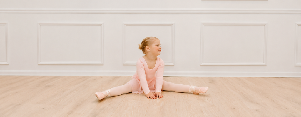 Toddler ballerina in a pink long-sleeve leotard, tights, and ballet shoes sitting in a split stretch on a light wood floor in a bright studio.

