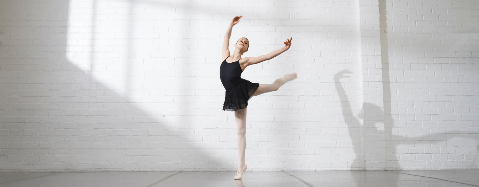 Young ballet dancer in a black leotard and skirt performing a leg extension against a white brick studio wall with soft window shadows.